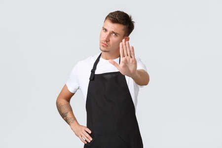 Employees, grocery stores and coffee shop concept. Serious-looking displeased barista, waiter in black apron extend hand for stop gesture, prohibit entering cafe, forbid action, white backgroundの写真素材