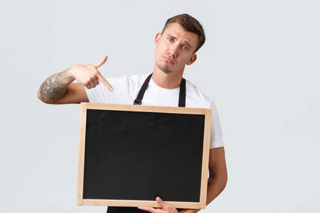 Small retail business owners, cafe and restaurant employees concept. Upset gloomy and distressed salesman pointing finger at chalk board without signs, standing white background disappointedの写真素材