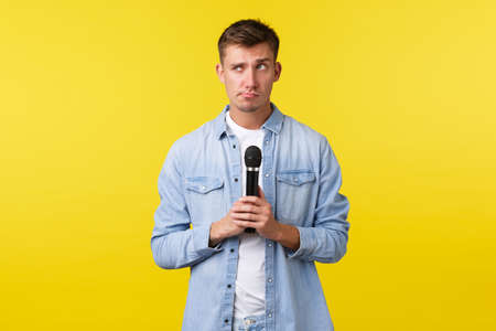 Lifestyle, people emotions and summer leisure concept. Indecisive thoughtful handsome man picking song, thinking what perform, holding microphone and looking away hesitant, yellow backgroundの写真素材