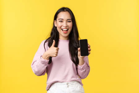 People emotions, lifestyle leisure and beauty concept. Smiling asian woman 20s, showing smartphone display, recommend application or mobile game, showing thumbs-up in approval, yellow backgroundの写真素材