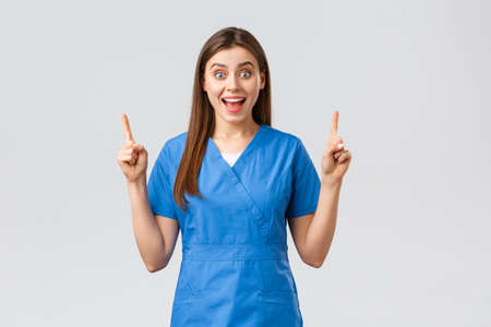 Healthcare workers, prevent virus, insurance and medicine concept. Excited, happy nurse or doctor in blue scrubs smiling from good news, pointing fingers up, show patients great newsの写真素材