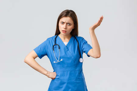 Medical workers, healthcare, covid-19 and vaccination concept. Frustrated and confused young female nurse, pretty doctor in blue scrubs, hear strange idea, shrug with hand raised in nonsenseの写真素材