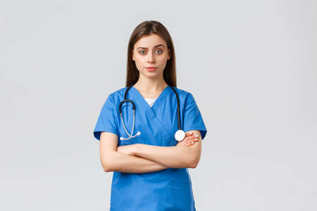 Healthcare workers, prevent virus, insurance and medicine concept. Skeptical female nurse or doctor in blue scrubs, cross arms chest and stare with impatients, smirk disapprovalの写真素材