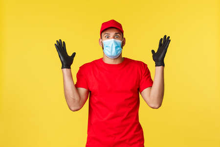 Express delivery during pandemic, covid-19, safe shipping, online shopping concept. Shocked and excited courier in uniform and medical mask, tell big news, stare impressed, yellow backgroundの写真素材