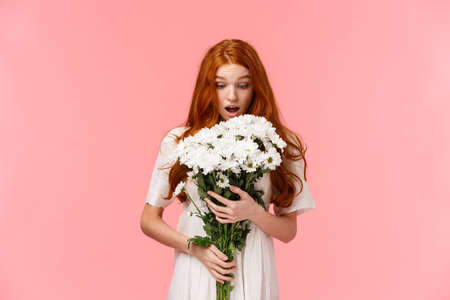 Surprised attractive redhead girl receive romantic valentines day gift, looking at beautiful bouquet of flowers amazed and wondered who might sender be, standing over pink backgroundの写真素材