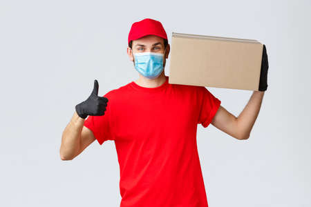 Packages and parcels delivery, covid-19 quarantine delivery, transfer orders. Cheerful courier in red uniform, gloves and face mask, thumb-up, no problem deliver order box to customersの写真素材