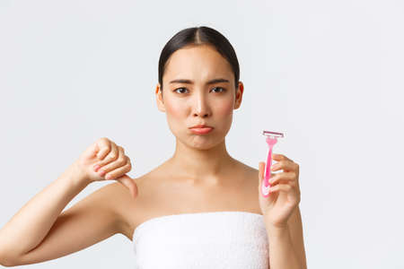 Beauty, personal care, and depilation concept. Close-up of disappointed asian girl in bath towel showing thumbs-down and razor, displeased with shaving quality, standing white backgroundの写真素材