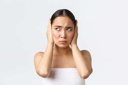 Beauty, cosmetology and spa salon concept. Bothered and disturbed young asian woman cant relax from loud noise, shut ears with hands and frowning, looking angry left, white backgroundの写真素材