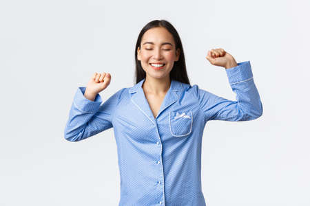 Smiling joyful asian girl in pajamas standing with closed eyes and delighted grin, stratching after good sleep, waking-up full of energy, getting ready to college, white backgroundの写真素材