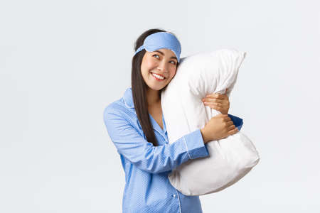 Dreamy beautiful asian girl in blue pajamas and sleeping mask hugging pillow and smiling pleased as looking upper left corner imaging something in bed, picturing, using imaginationの写真素材
