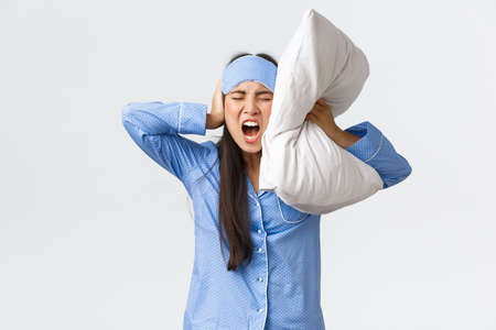 Angry furious asian girl in pajamas and sleeping mask, lying bed and shut ears with pillow, shouting mad as cant sleep from loud noise, neighbours through party at night, complaining annoying soundの写真素材