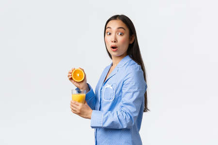 Morning, active and healthy lifestyle and home concept. Surprised asian girl caught on making orange juice, looking in awe with dropped jaw at camera, stand in pajamas with glass and half of orangeの写真素材