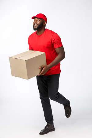 Portrait of Happy African American delivery man in red cloth walking to send a box package to customer. Isolated on Grey studio Background. Copy Spaceの写真素材
