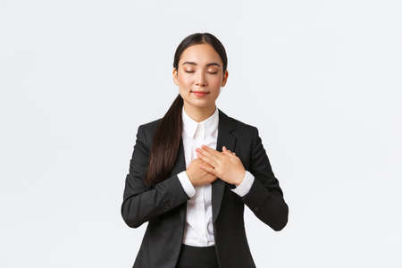 Happy dreamy asian female entrepreneur in suit close eyes and holding hands on heart, remember lovely memory, thinking about something pleasant over white backgroundの写真素材