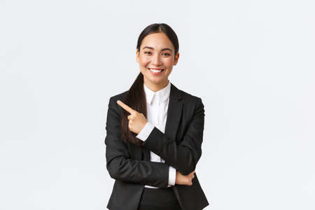 Happy professional asian female manager, businesswoman in suit showing announcement, smiling and pointing finger left at product or project banner, standing white backgroundの写真素材