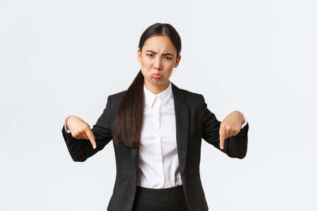 Disappointed sulking asian businesswoman complaining on unfair situation. Female entrepreneur in suit pointing fingers down and grimacing displeased, standing upset white backgroundの写真素材
