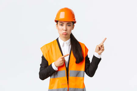 Building, construction and industrial concept. Upset and disappointed female asian manager, engineer in safety helmet pouting, complaining something bad, pointing fingers upper right cornerの写真素材