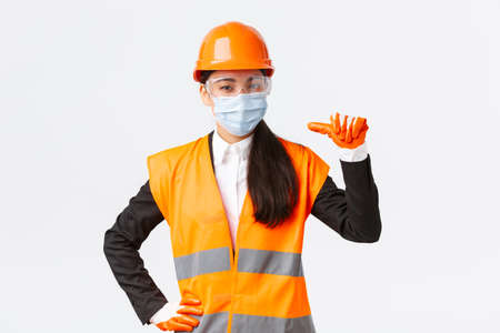 Covid-19 safety protocol at enterpise, construction and preventing virus concept. Confident asian female engineer in helmet and face mask pointing at herself, advertise personal assitanceの写真素材