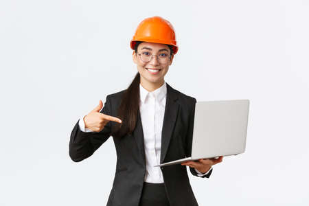 Smiling pleased asian female chief engineer showing chart with enterprise profit, making presentation during investors meeting, wearing safety helmet, pointing at laptop screen, white backgroundの写真素材
