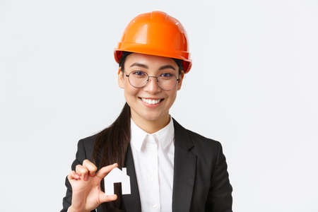 Close-up of happy asian female engingeer, real estate agent in helmet and business suit holding house miniature and smiling, architect working over renovation project, white backgroundの写真素材