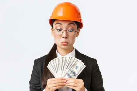 Impressed and pleased female asian construction inspector, industrial architect receiving big bribe, counting money and looking astonished with sum, standing in safety helmet and suitの写真素材
