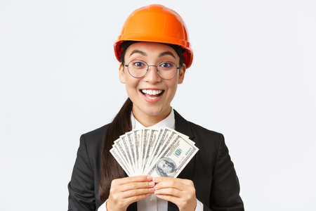 Close-up of happy and excited female asian entrepreneur at factory, wear safety helmet and business suit, showing big income, holding money and rejoicing, smiling pleased, white backgroundの写真素材