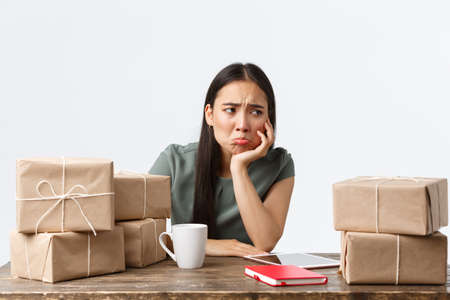 Small business owners, startup and e-commerce concept. Sad and gloomy asian businesswoman pouting as not have clients, waiting for orders, looking distressed sitting near piles of boxesの写真素材