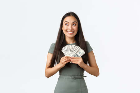 Small business owners, women entrepreneurs concept. Dreamy excited, successful winning woman in dress thinking how invest or what buy while holding big sum of money, standing white backgroundの写真素材