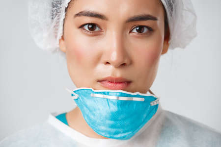 Covid-19, coronavirus disease, healthcare workers concept. Close-up of exhausted serious-looking asian female doctor take-off personal protective equipment, have skin damage from respiratorの写真素材