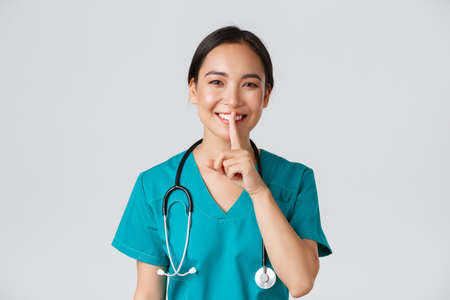 Healthcare workers, preventing virus, quarantine campaign concept. Smiling pretty asian doctor, nurse in scrubs smiling, shushing, asking stay quiet, standing white backgroundの写真素材