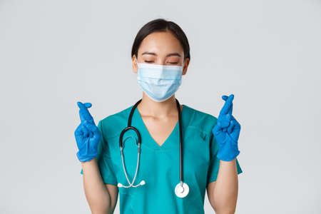 Covid-19, coronavirus disease, healthcare workers concept. Close-up of hopeful asian female doctor, physician in medical mask and rubber gloves, making wish, cross fingers good luckの写真素材