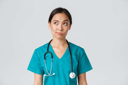 Healthcare workers, preventing virus, quarantine campaign concept. Thoughtful asian female doctor, nurse in scrubs looking upper left corner and smirk unsure, thinking, white backgroundの写真素材