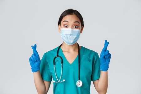Covid-19, coronavirus disease, healthcare workers concept. Excited and hopeful asian female doctor, nurse in medical mask and rubber gloves cross fingers good luck, makign wish, white backgroundの写真素材