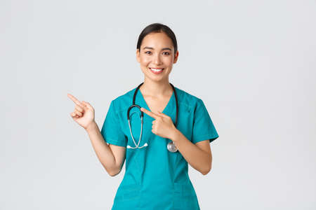 Covid-19, healthcare workers, pandemic concept. Smiling pretty asian doctor, nurse in scrubs and stethoscope, pointing fingers upper left corner, showing online clinic banner, white backgroundの写真素材