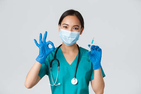 Covid-19, coronavirus disease, healthcare workers concept. Cute asian female doctor, physician in medical mask and rubber gloves show okay gesture and hold syringe with vaccine, white backgroundの写真素材