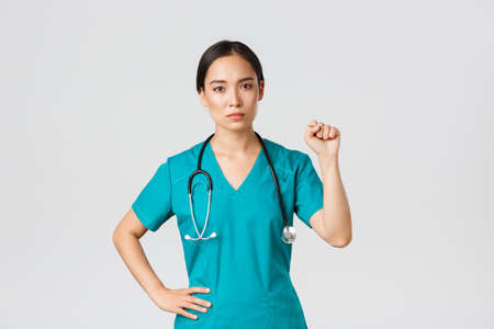 Covid-19, healthcare workers, pandemic concept. Serious-looking confident serious asian female doctor showing support to fellow collegues during coronavirus, raising fist in unity gestureの写真素材
