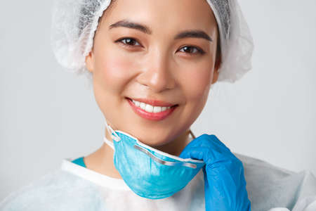 Covid-19, coronavirus disease, healthcare workers concept. Close-up of hopeful happy smiling asian female doctor in personal protective equipment, take-off respirator and have skin damageの写真素材