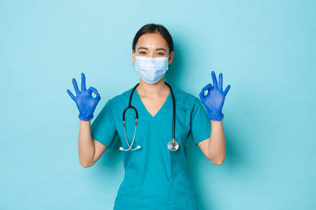 Covid-19, social distancing and coronavirus pandemic concept. Satisfied smiling asian female doctor, nurse in medical mask and scrubs, rubber gloves, ensure everything alright, show okay gestureの写真素材
