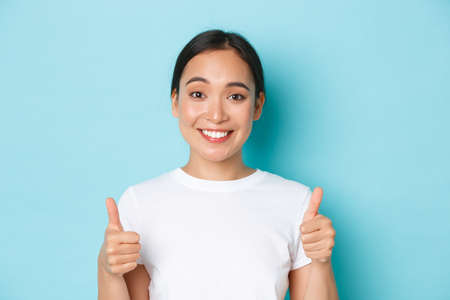 Close-up of satisfied and cheerful smiling asian girl in white t-shirt, showing thumbs-up supportive and looking pleased at camera, recommend product, compliment good choiceの写真素材