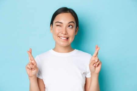 Close-up of hopeful dreamy asian girl making wish, looking upper left corner thoughtful, standing light blue background with fingers crossed for good luck, praying or anticipating positive newsの写真素材