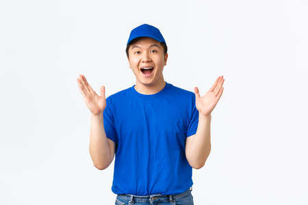 Happy and impressed asian delivery guy in blue courier clap hands, applause glad, praising nice work. Cheerful courier celebrating big event in company, achieve goal, white backgroundの写真素材