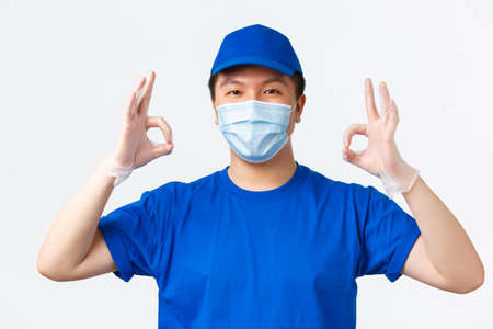 Online shopping, delivery and coronavirus pandemic, preventing virus concept. Confident smiling asian courier in blue uniform, medical mask and gloves showing okay gesture, provide excellent serviceの写真素材