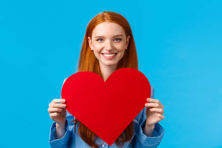 Happy, cheerful and lovely, tender caucasian woman holding big red heart valentines card and smiling, giving her love and care, congratulate boyfriend with their romantic date, blue backgroundの写真素材