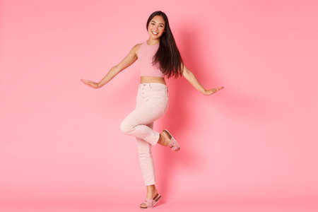 Fashion, beauty and lifestyle concept. Silly and glamour beautiful asian girl posing coquettish over pink background, smiling and raising foot, advertisement of women selfcare and shoppingの写真素材