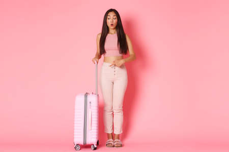 Travelling, holidays and vacation concept. Full-length of surprised and curious glamour asian girl in summer clothes, standing with suitcase, pointing lower left corner, pink backgroundの写真素材