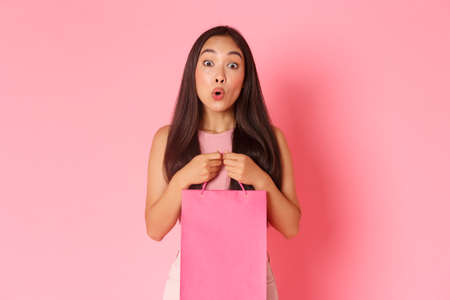 Beauty, fashion and lifestyle concept. Gorgeous happy asian girl receive gift, holding shopping bag and smiling dreamy, looking thankful, standing over pink backgroundの写真素材