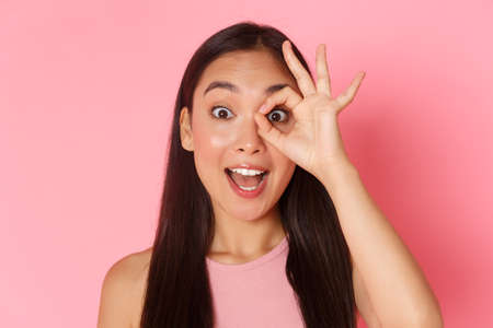 Beauty, fashion and lifestyle concept. Close-up of impressed beautiful asian girl see something amazing, stare at camera amazed, looking through okay gesture and say wow, pink backgroundの写真素材