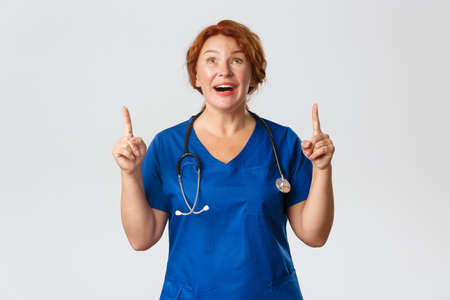 Medicine, healthcare and coronavirus concept. Amazed and fascinated middle-aged redhead female doctor, nurse in scrubs looking and pointing fingers up with admiration, grey backgroundの写真素材