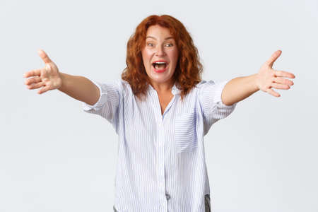 Excited and happy redhead middle-aged woman inviting to come in, extending hands forward for hug, embracing family, greeting someone with friendly attitude, white backgroundの写真素材