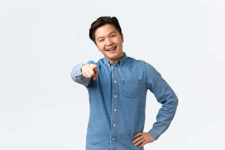 Smiling happy asian handsome guy in blue shirt, have braces, pointing finger at camera, encourage person click link, naming or choosing someone, standing over white backgroundの写真素材